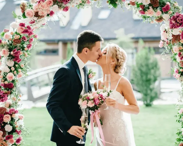 Novios besándose bajo un arco floral rosa y blanco, celebrando su boda barata con copas de champán.