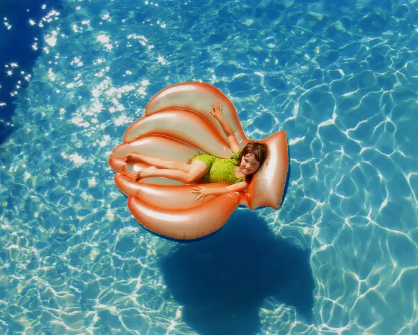 Niña sonriente en un flotador naranja con forma de concha, disfrutando de la pool party y sus juegos bajo el sol.