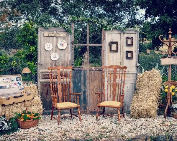 Un rústico photocall boda casero con puertas antiguas, sillas de madera, balas de heno y detalles florales.