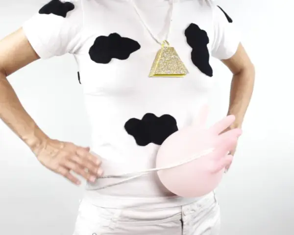Persona con un disfraz vaca casero: camiseta blanca con manchas negras, collar de campana dorada y un globo rosa simulando la ubre.