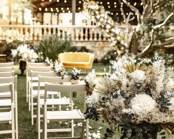 Sillas blancas alineadas en un jardín, un sofá amarillo y un gran arreglo floral. Ideas para como hacer una boda económica y hermosa.