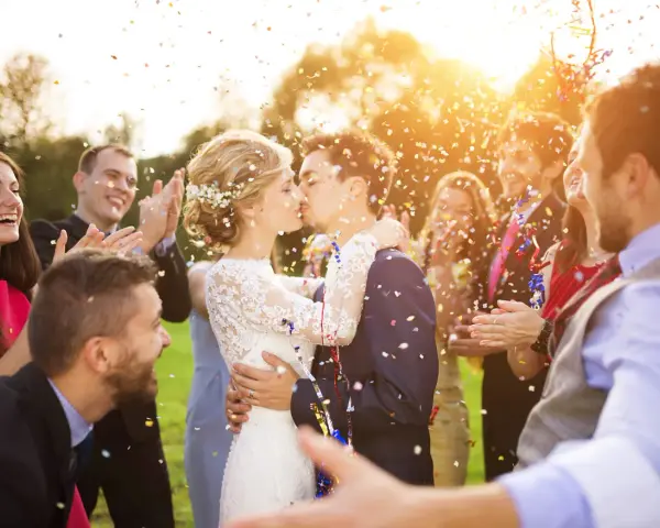 Novios besándose entre confeti mientras amigos aplauden y ríen. ¡Un momento perfecto para felicitar a unos novios!