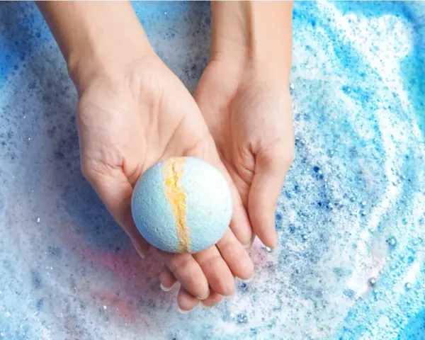 Manos sostienen una bomba de baño azul y amarilla sobre espuma burbujeante. ¡Perfecto para hacer bombas de baño caseras!