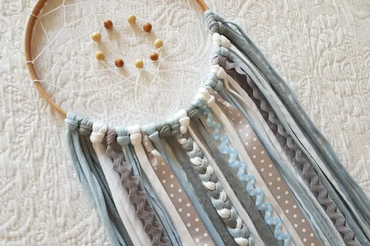 Un atrapasueños casero con aros de madera, red blanca y cuentas. Cintas de tela y encaje cuelgan en tonos grises, blancos y azules.
