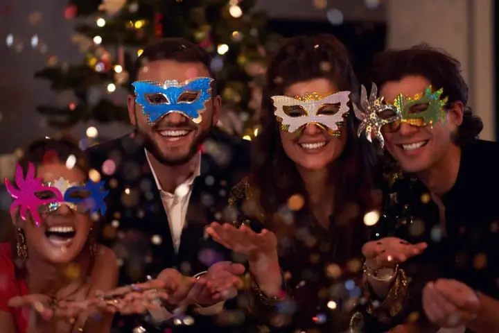Cuatro amigos disfrutan de una temática fiesta de mascaras, riendo y lanzando confeti frente a un árbol de Navidad iluminado.