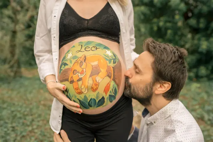 Barriga de embarazada pintada con un león y el nombre "Leo". El padre besa la barriga. Unas plantillas para pintar barriga de embarazada niño, como esta, son un recuerdo precioso.