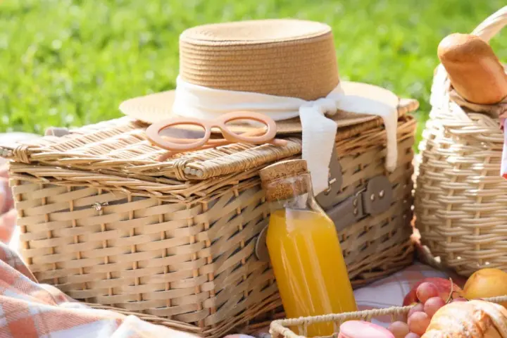 Cajas picnic listas para disfrutar: sombrero, gafas de sol, jugo y frutas sobre mantas a cuadros en un día soleado.
