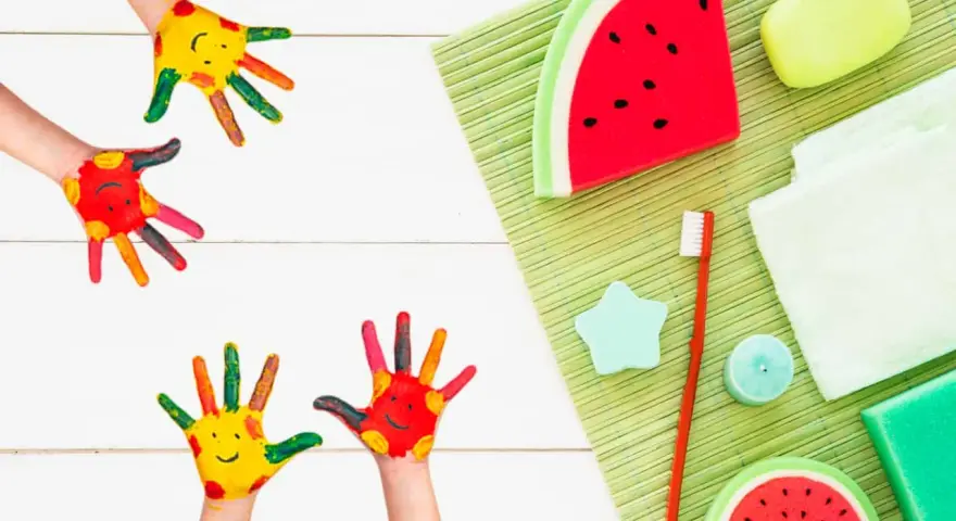 Manos pintadas de colores con caritas sonrientes, listas para hacer jabones para niños. Hay jabones con forma de sandía y otros accesorios de baño.
