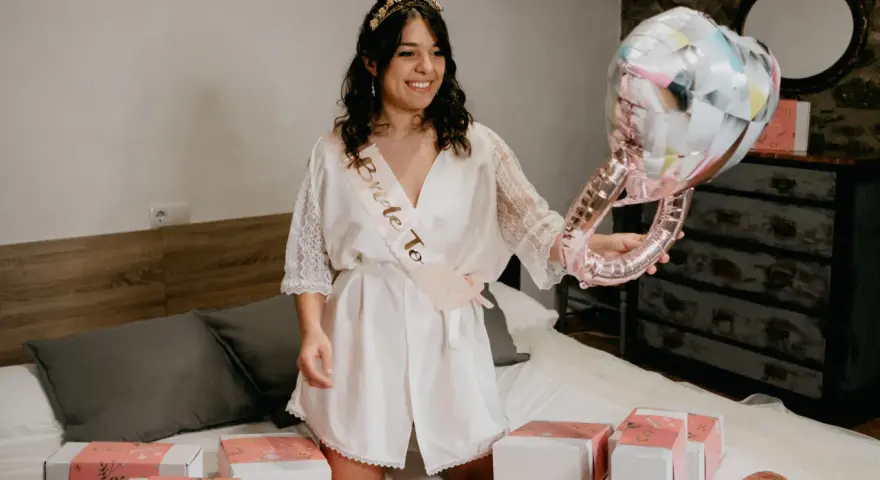 Novia sonriente con corona y banda "Bride To Be", sosteniendo un globo de anillo. Alrededor, cajas de regalo y un pack de accesorios para despedida de soltera.