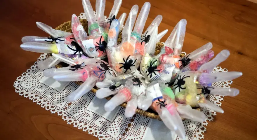Manualidades chuches Halloween: guantes transparentes rellenos de caramelos y decorados con arañas negras, presentados en una cesta sobre un mantel de encaje.