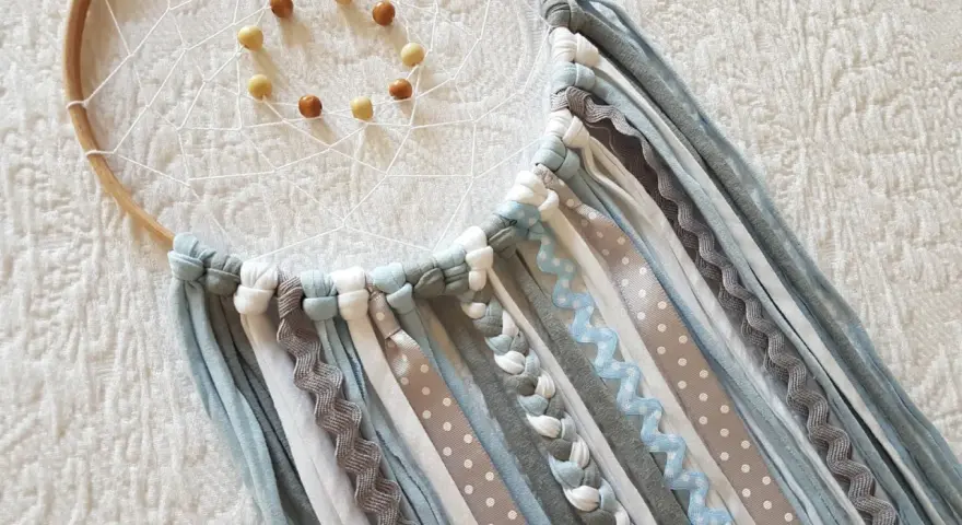 Un atrapasueños casero con aros de madera, red blanca y cuentas. Cintas de tela y encaje cuelgan en tonos grises, blancos y azules.