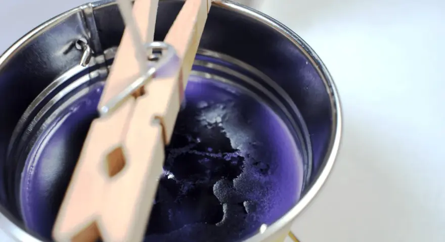 Proceso para como hacer velas de soja: cera morada enfriándose en un recipiente metálico, sujeta por pinzas de madera.