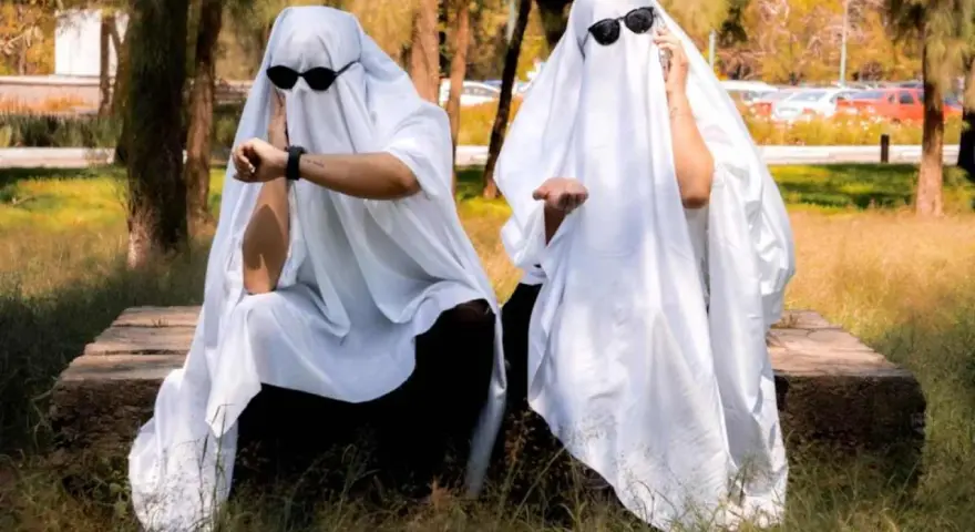 Dos figuras cubiertas con sábanas blancas y gafas de sol, sentadas en un banco de piedra. Una mira su reloj, la otra extiende la mano. ¡Ideas de terror para un disfraz!