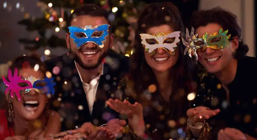 Cuatro amigos disfrutan de una temática fiesta de mascaras, riendo y lanzando confeti frente a un árbol de Navidad iluminado.