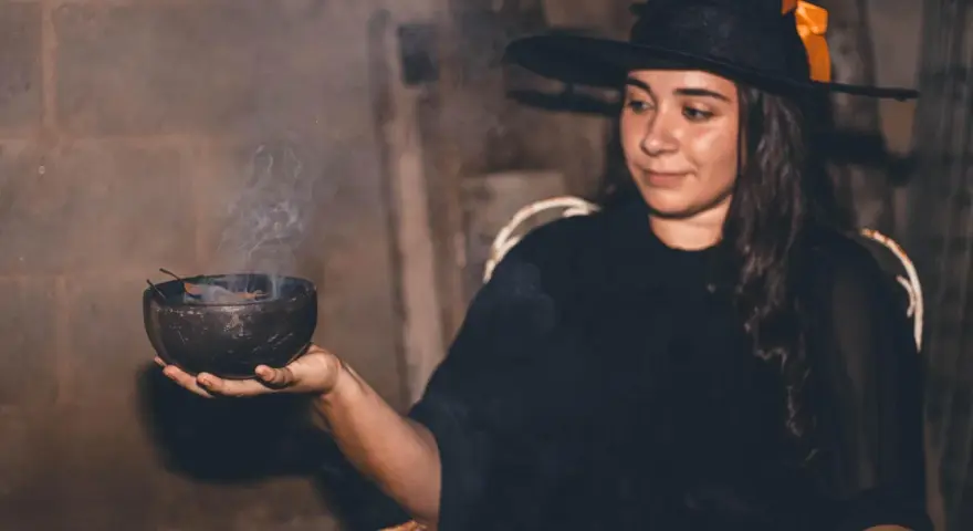 Mujer con disfraz bruja casero adulto, con sombrero negro y naranja, sosteniendo un cuenco humeante.