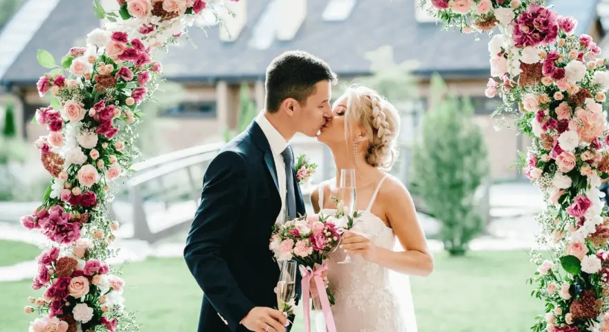 Novios besándose bajo un arco floral rosa y blanco, celebrando su boda barata con copas de champán.