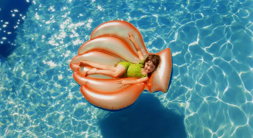Niña sonriente en un flotador naranja con forma de concha, disfrutando de la pool party y sus juegos bajo el sol.