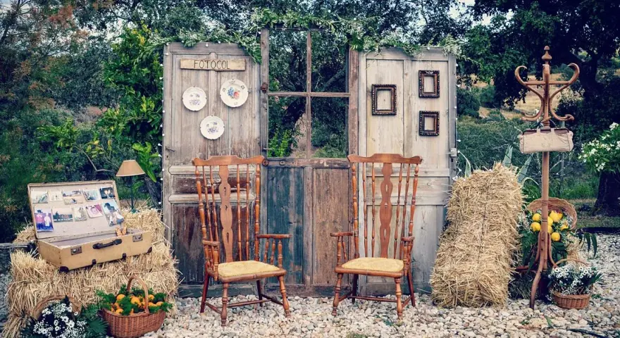 Un rústico photocall boda casero con puertas antiguas, sillas de madera, balas de heno y detalles florales.