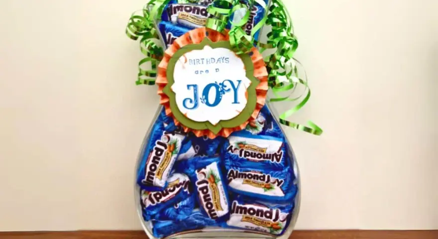Un jarrón lleno de botes de chuches caseros, decorado con un lazo verde y un mensaje que dice "BIRTHDAYS are a JOY".