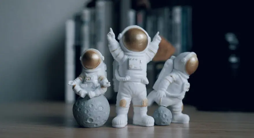 Tres figuras de un disfraz astronauta casero, una meditando sobre la luna, otra celebrando y la tercera jugando con una pequeña luna.