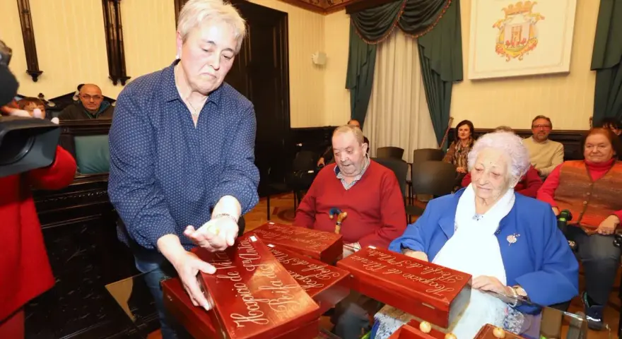 Mujer saca bolas de una caja de madera, pensando en ideas para sortear en una rifa. Personas observan.