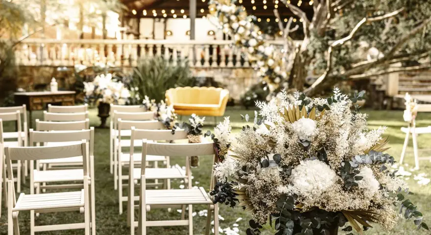 Sillas blancas alineadas en un jardín, un sofá amarillo y un gran arreglo floral. Ideas para como hacer una boda económica y hermosa.
