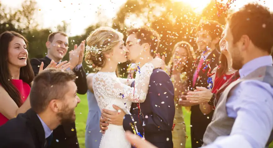 Novios besándose entre confeti mientras amigos aplauden y ríen. ¡Un momento perfecto para felicitar a unos novios!