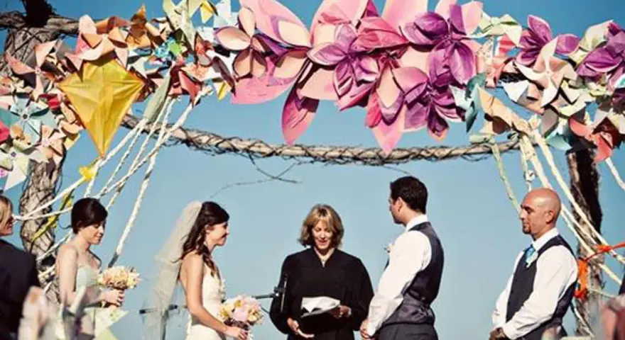 Boda al aire libre con un arco decorado con manualidades de papel coloridas. La pareja se da el sí ante un oficiante.