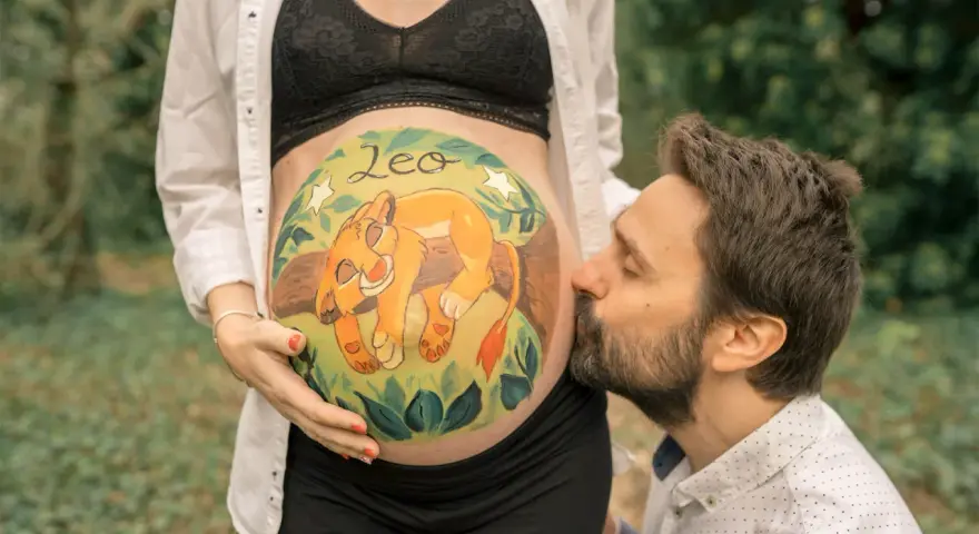 Barriga de embarazada pintada con un león y el nombre "Leo". El padre besa la barriga. Unas plantillas para pintar barriga de embarazada niño, como esta, son un recuerdo precioso.