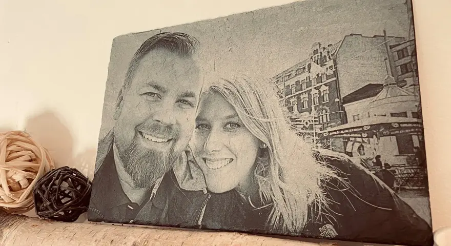 Foto piedra con retrato de pareja sonriente, con edificios y un puesto de helados al fondo.