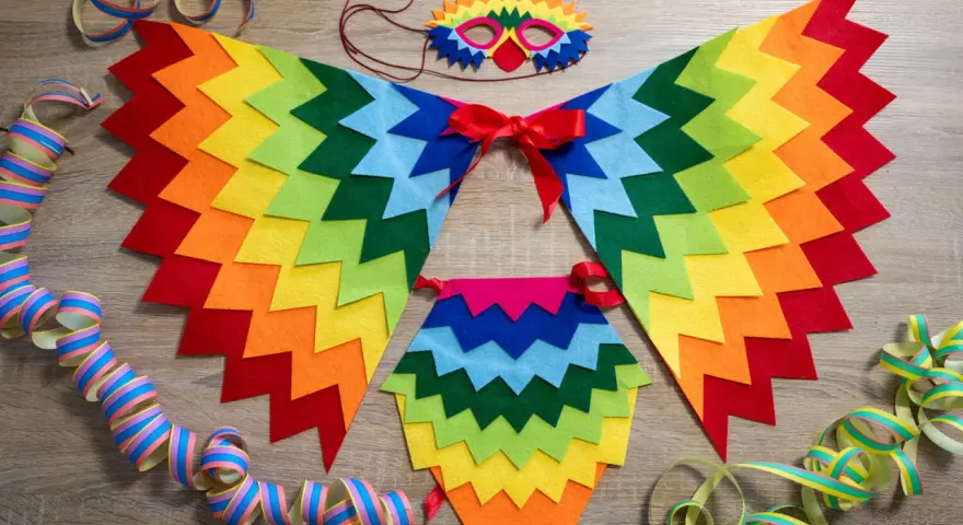 Un colorido disfraz de pájaro casero con alas de fieltro, máscara y adornos de serpentinas, listo para una fiesta.