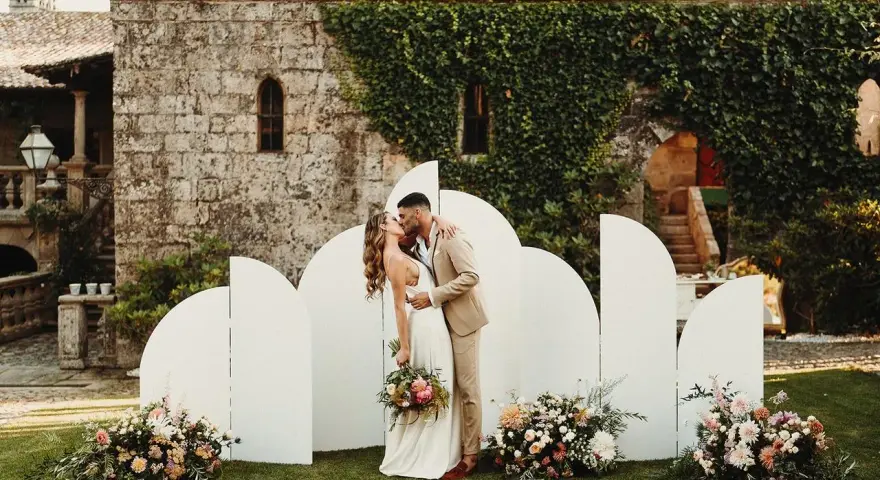 Pareja besándose frente a un photocall bodas de oro casero, decorado con flores y un fondo arquitectónico.