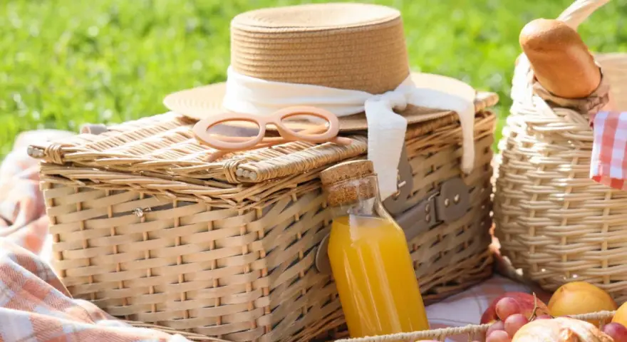 Cajas picnic listas para disfrutar: sombrero, gafas de sol, jugo y frutas sobre mantas a cuadros en un día soleado.