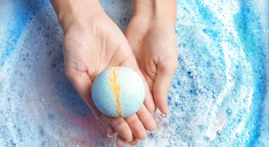 Manos sostienen una bomba de baño azul y amarilla sobre espuma burbujeante. ¡Perfecto para hacer bombas de baño caseras!