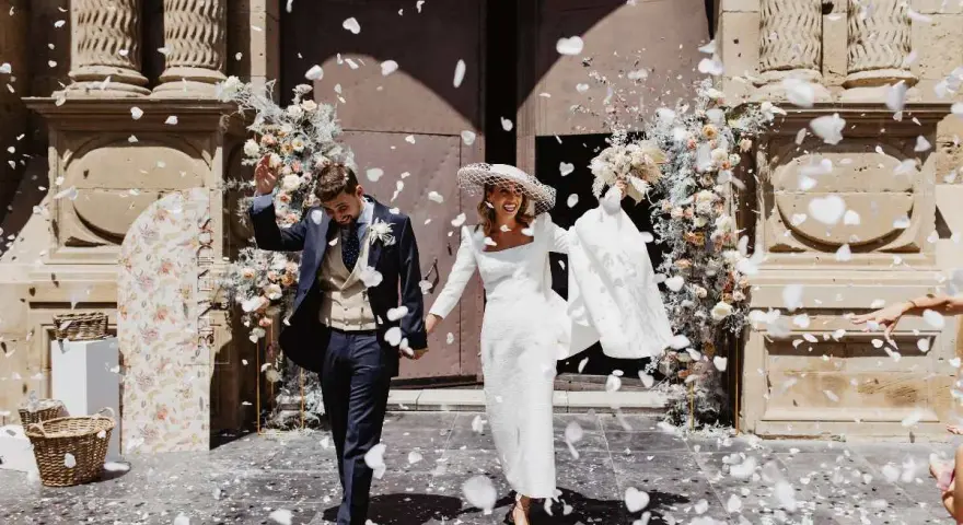 Novios felices salen de la iglesia, cubiertos de pétalos blancos. Es un momento perfecto para tirar confeti en una boda.