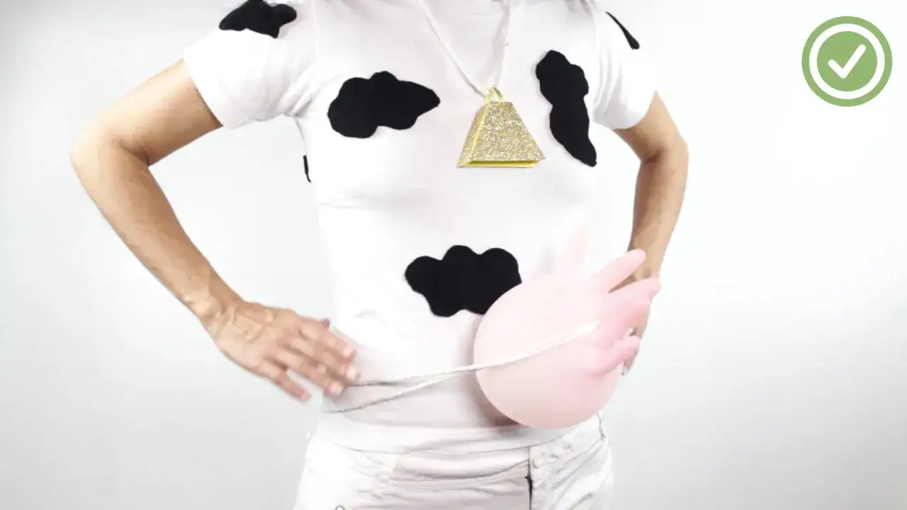 Persona con un disfraz vaca casero: camiseta blanca con manchas negras, collar de campana dorada y un globo rosa simulando la ubre.
