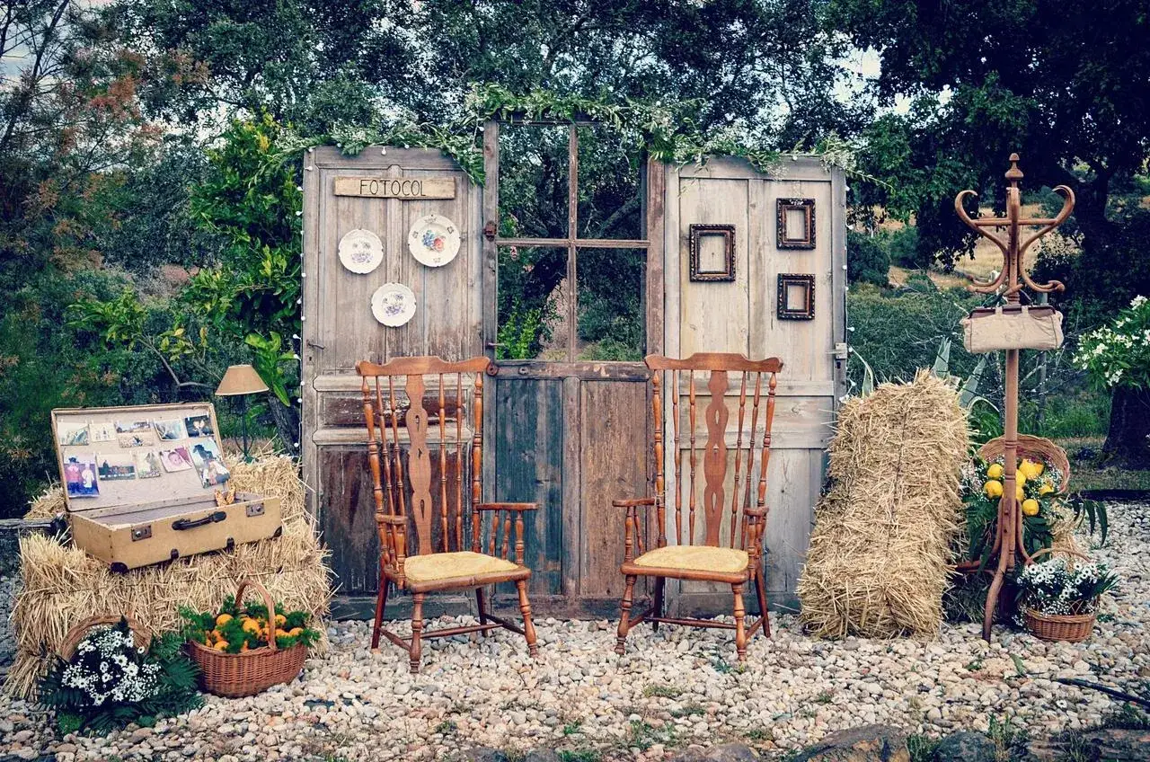 Un photocall bodas de oro casero con puertas rústicas, sillas de madera, balas de heno y un baúl lleno de recuerdos.