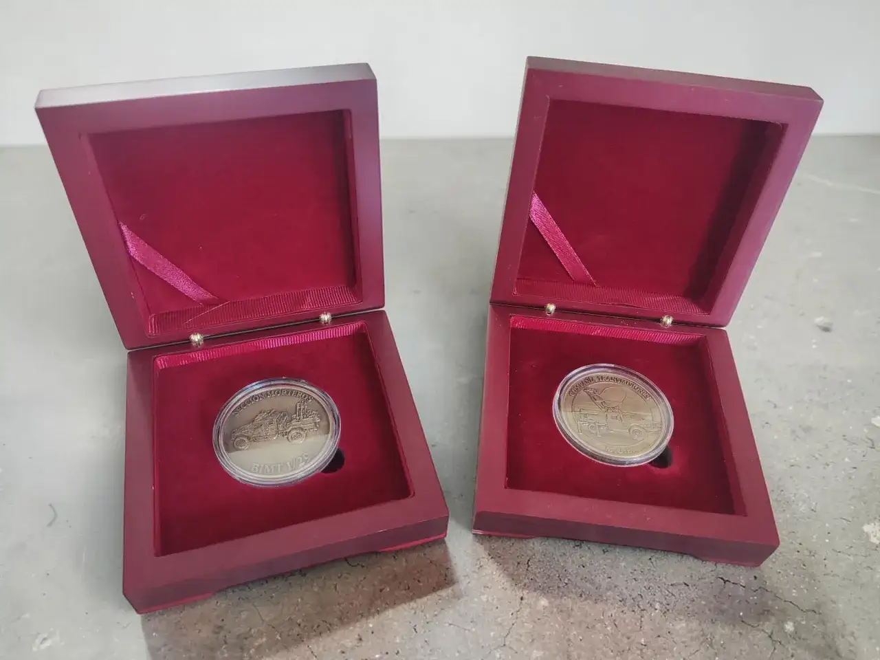 Dos cajas abiertas de terciopelo rojo exhiben dos monedas personalizadas con grabados de veh&iacute;culos.