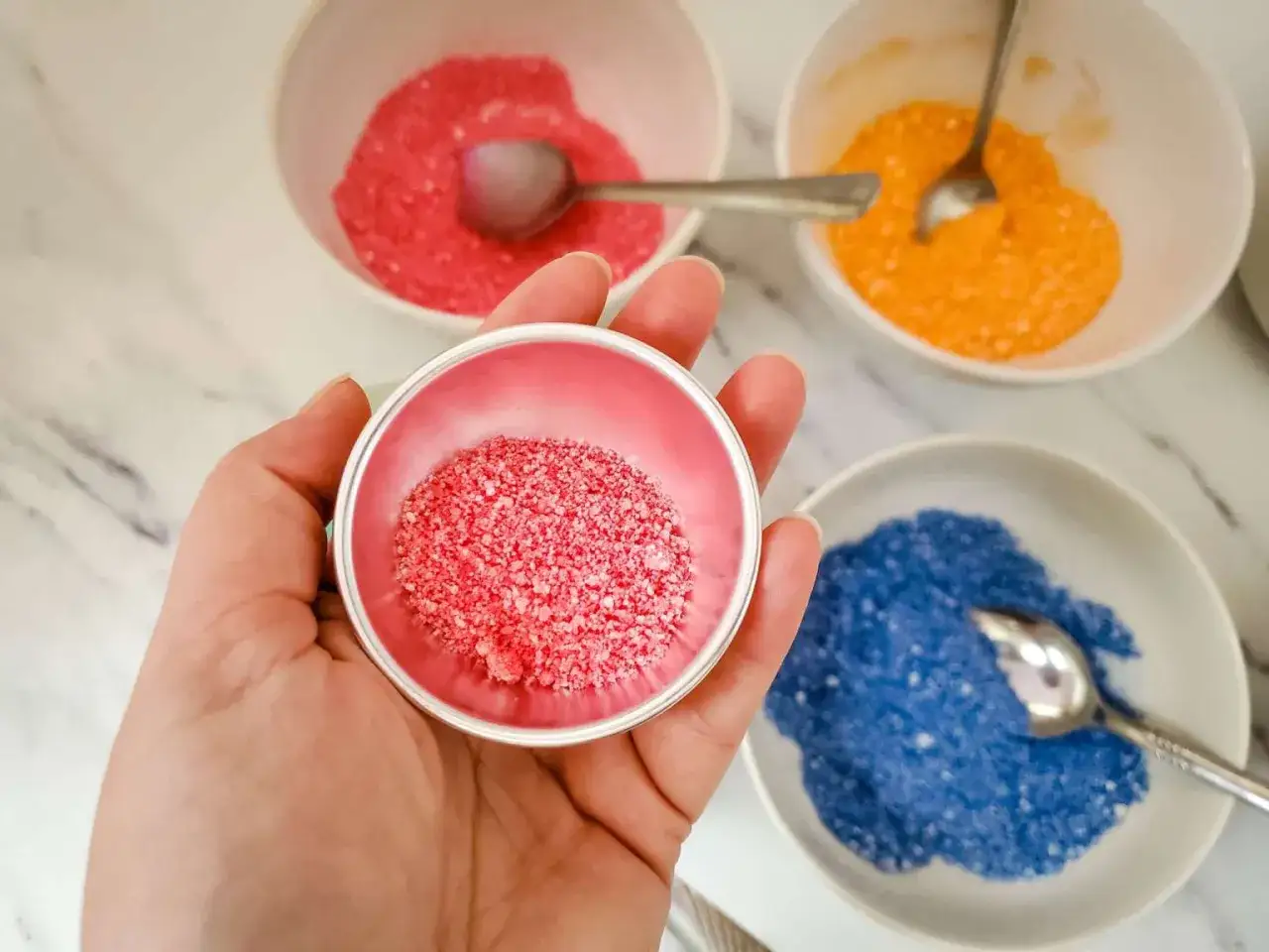 Mano sostiene un cuenco con polvo rosa, listo para hacer bombas de ba&ntilde;o. Otros cuencos con polvos de colores esperan.