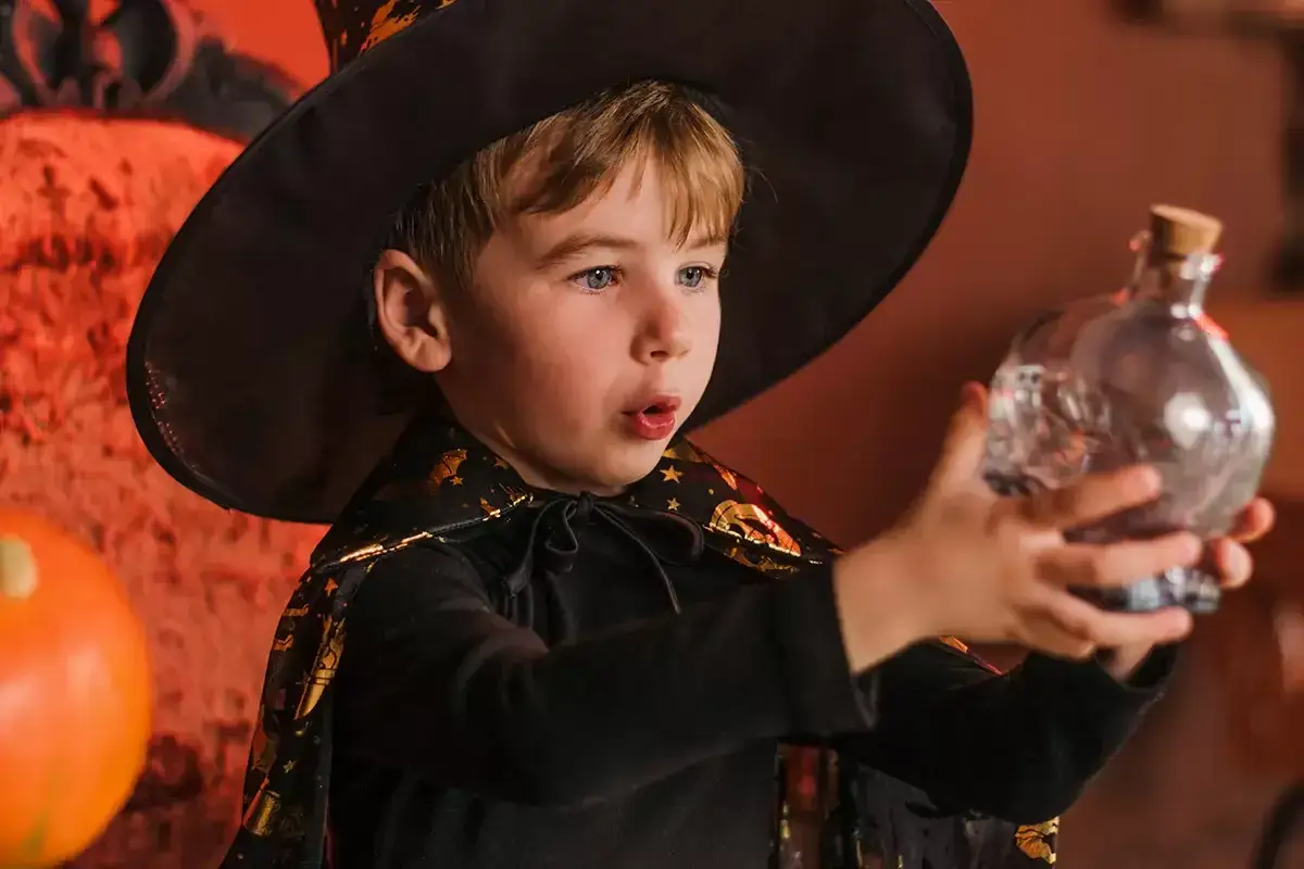 Niño mago con sombrero y capa, practicando trucos de magia para niños con las manos, sosteniendo una botella con forma de calavera.