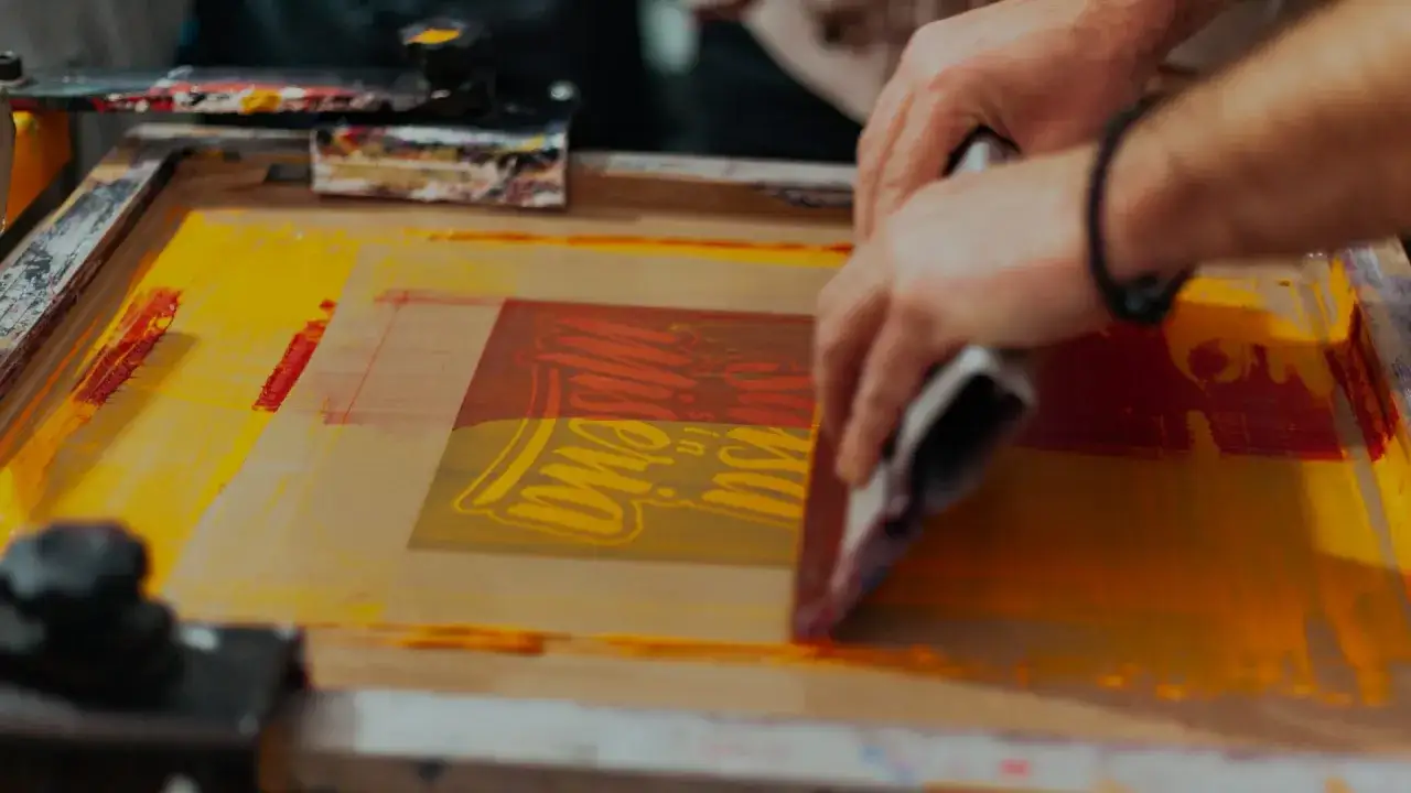 Manos aplicando tinta amarilla y roja sobre un diseño con serigrafía que es una técnica de impresión.