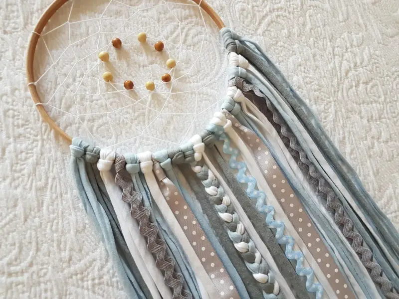 Un atrapasueños casero con aros de madera, red blanca y cuentas. Cintas de tela y encaje cuelgan en tonos grises, blancos y azules.