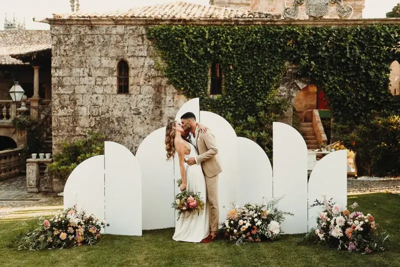 Pareja besándose frente a un photocall bodas de oro casero, decorado con flores y un fondo arquitectónico.