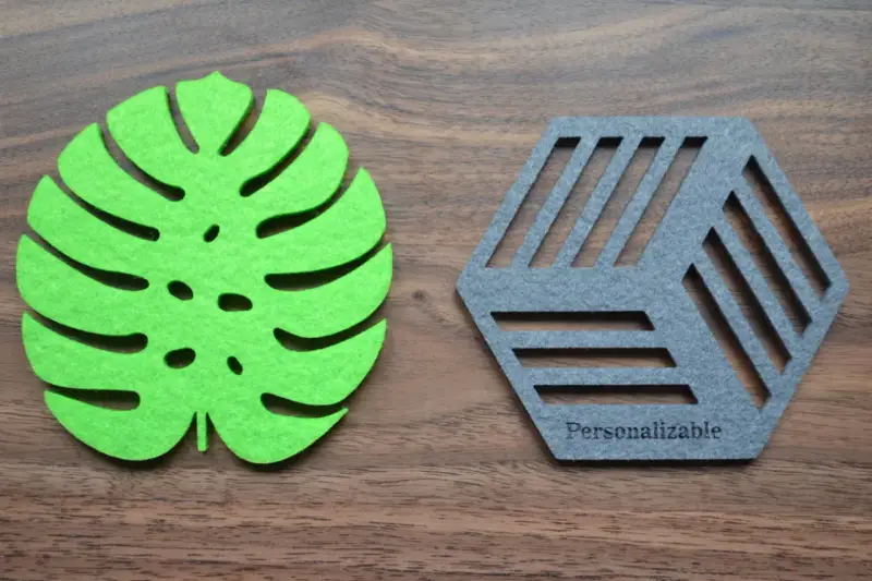 Posavasos de fieltro verde con forma de hoja y otro gris hexagonal con líneas, sobre fondo de madera.